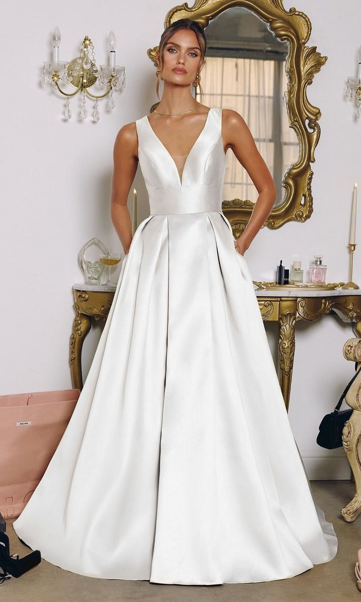 A woman wears the Long White Prom Dress: Amoris T814W in off white, featuring a sleeveless satin A-line design with a deep V-neckline, as she stands before an ornate gold mirror and vanity table.